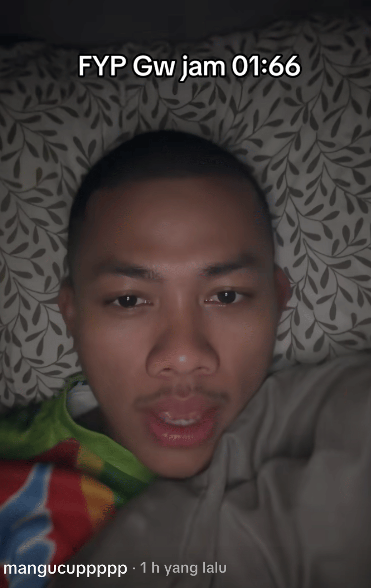 Seorang pria berbaring di tempat tidur dengan selimut abu-abu dan sarung bantal bermotif daun, menatap kamera dengan teks FYP Gw jam 01:66 di atasnya.