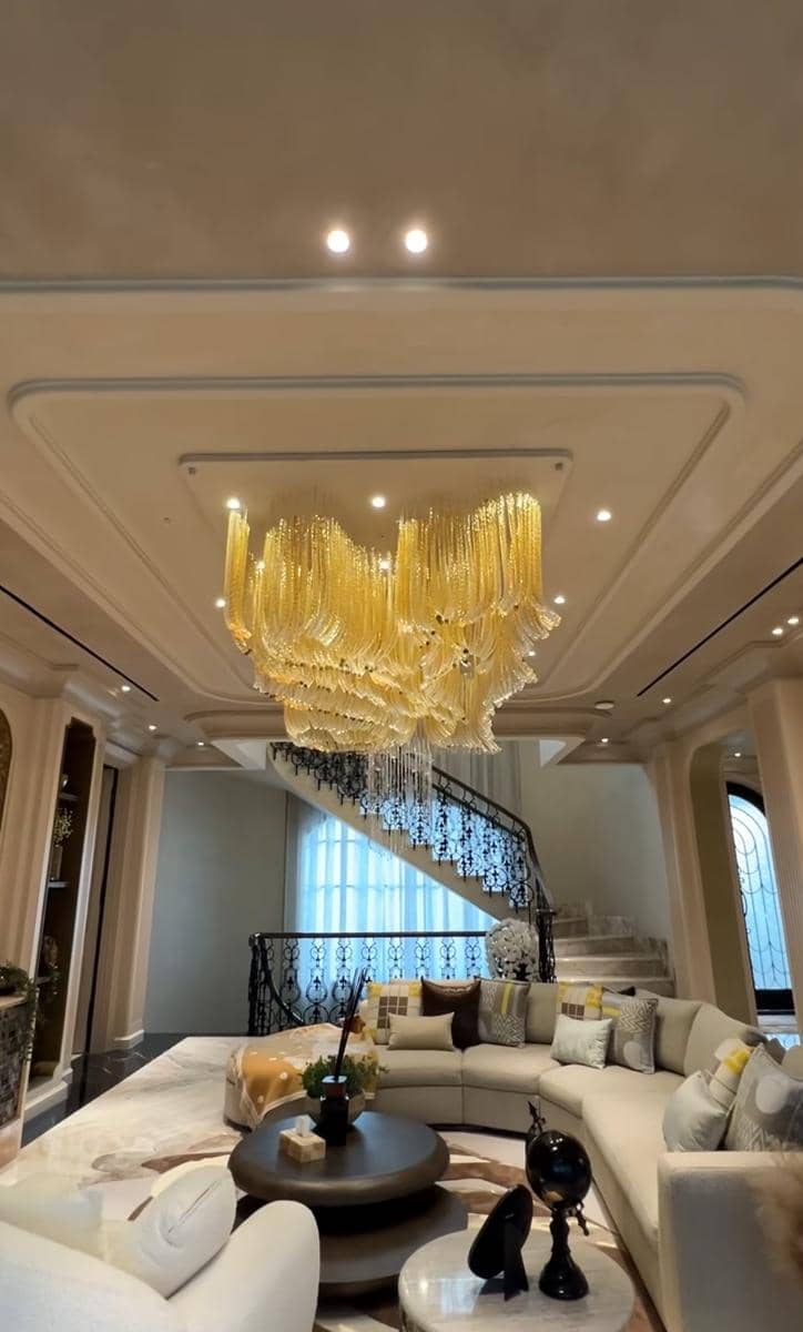Ruang tamu mewah dengan lampu gantung kristal besar berwarna emas, tangga melingkar berpagar besi, dan sofa melingkar elegan.