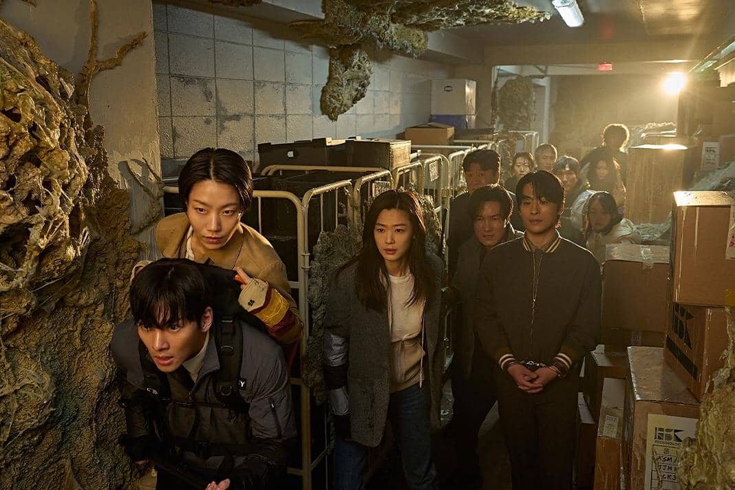 Sekelompok orang berjalan hati-hati di lorong sempit penuh kotak dan dinding berlumut dalam adegan film Korea Colony.