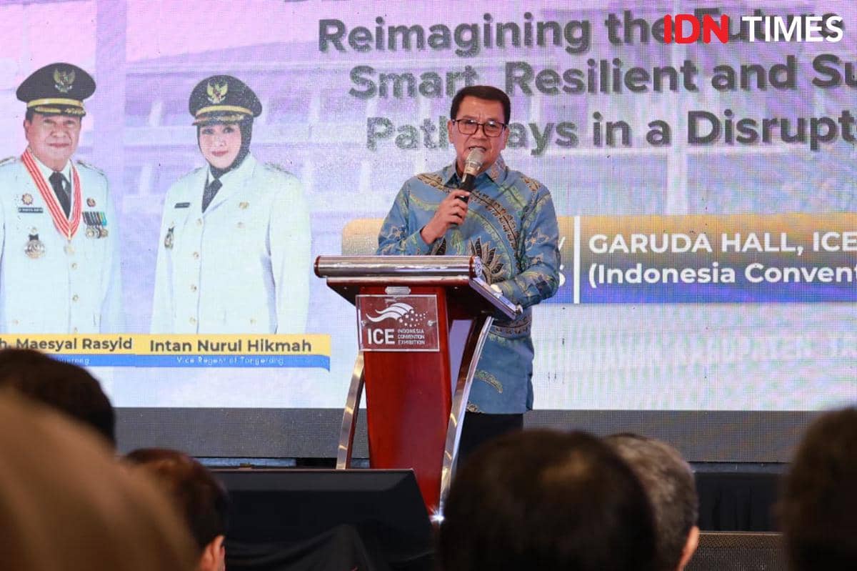 Seorang pria berbicara di podium dengan latar belakang layar besar berisi tulisan dan foto dua pejabat berpakaian dinas putih.