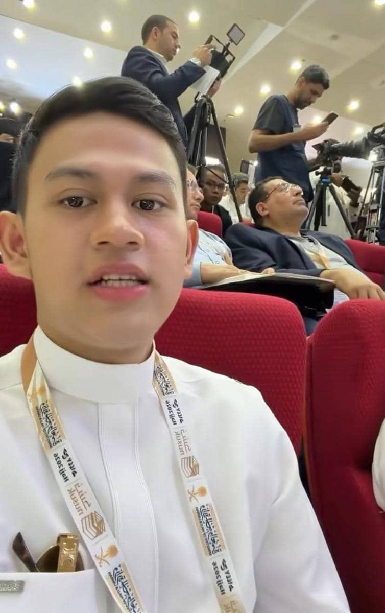 Seorang pria muda mengenakan pakaian putih dan lanyard duduk di kursi auditorium merah bersama peserta lain dalam sebuah acara.