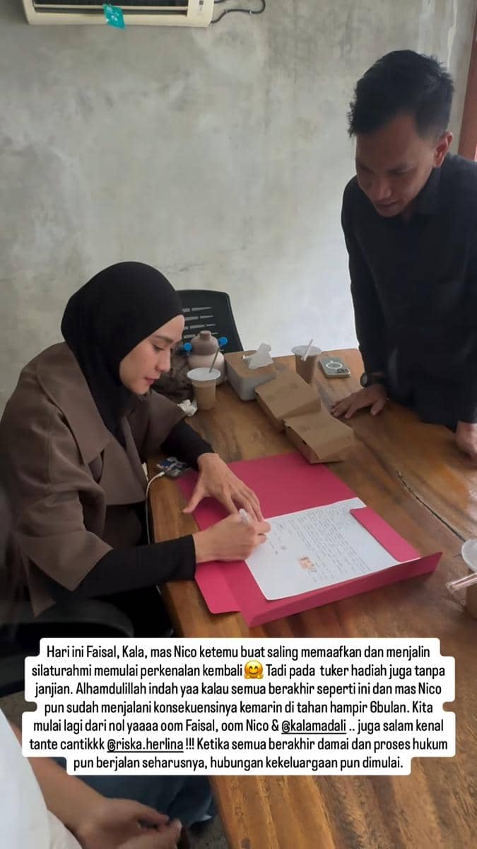 Seorang perempuan berhijab menandatangani dokumen di atas meja kayu dengan seorang pria berdiri di sampingnya di sebuah ruangan sederhana.