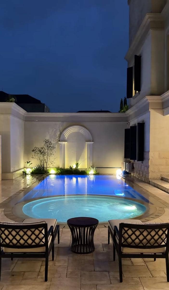 Kolam renang rumah mewah dengan pencahayaan malam berwarna biru dan area duduk elegan di tepi kolam.