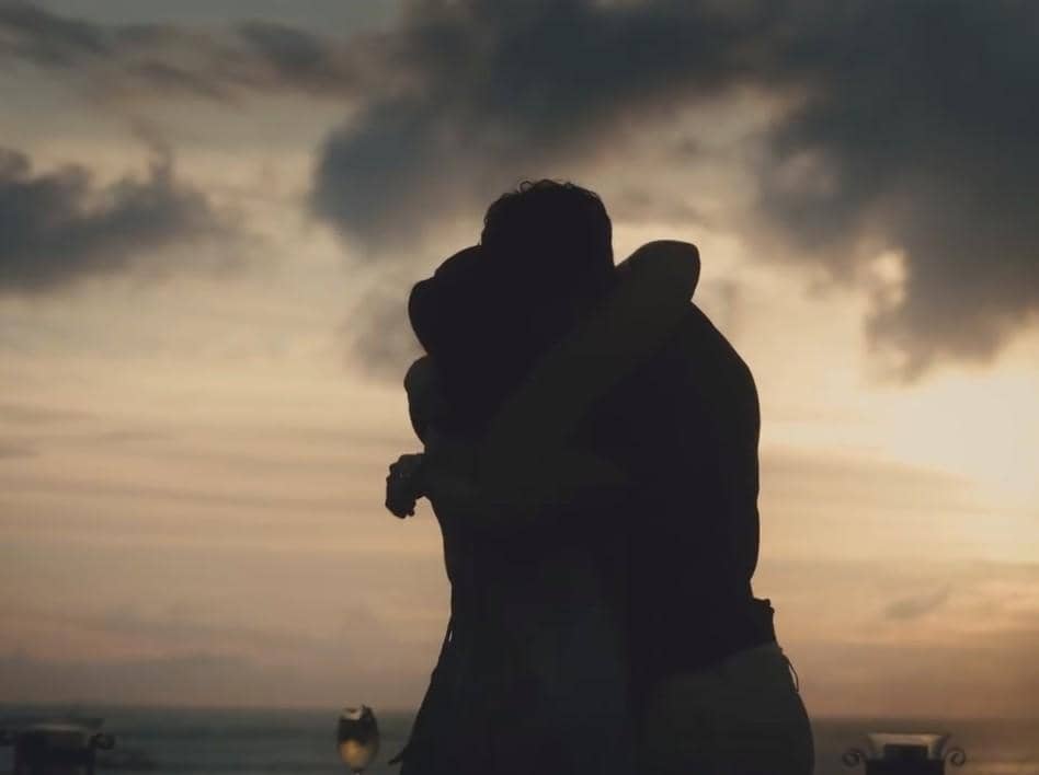 Sepasang kekasih berpelukan di tepi pantai saat matahari terbenam dengan latar langit berawan yang indah dan suasana romantis.