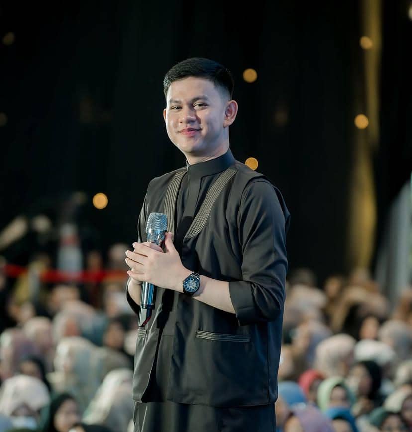 Seorang pria mengenakan pakaian hitam memegang mikrofon di atas panggung dengan latar penonton yang ramai dan pencahayaan lembut.