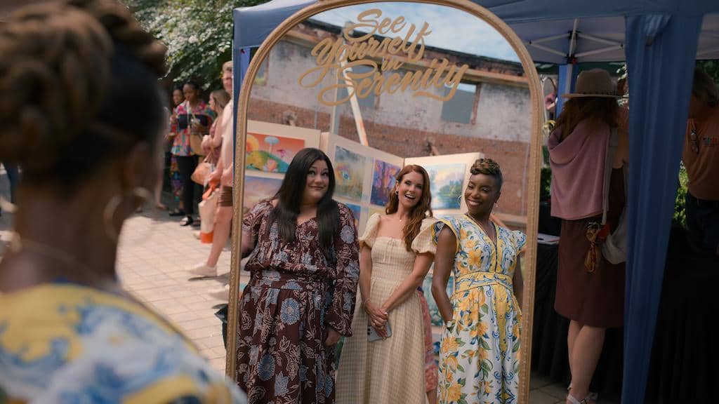 Tiga wanita tersenyum berdiri di depan cermin bertuliskan 'See Yourself in Serenity' dalam adegan Sweet Magnolias Season 5.