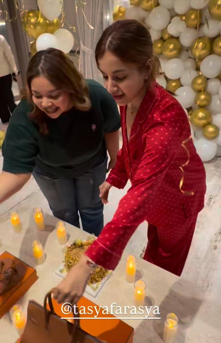 Dua wanita tersenyum saat merayakan ulang tahun dengan dekorasi balon putih dan emas serta lilin di atas meja.