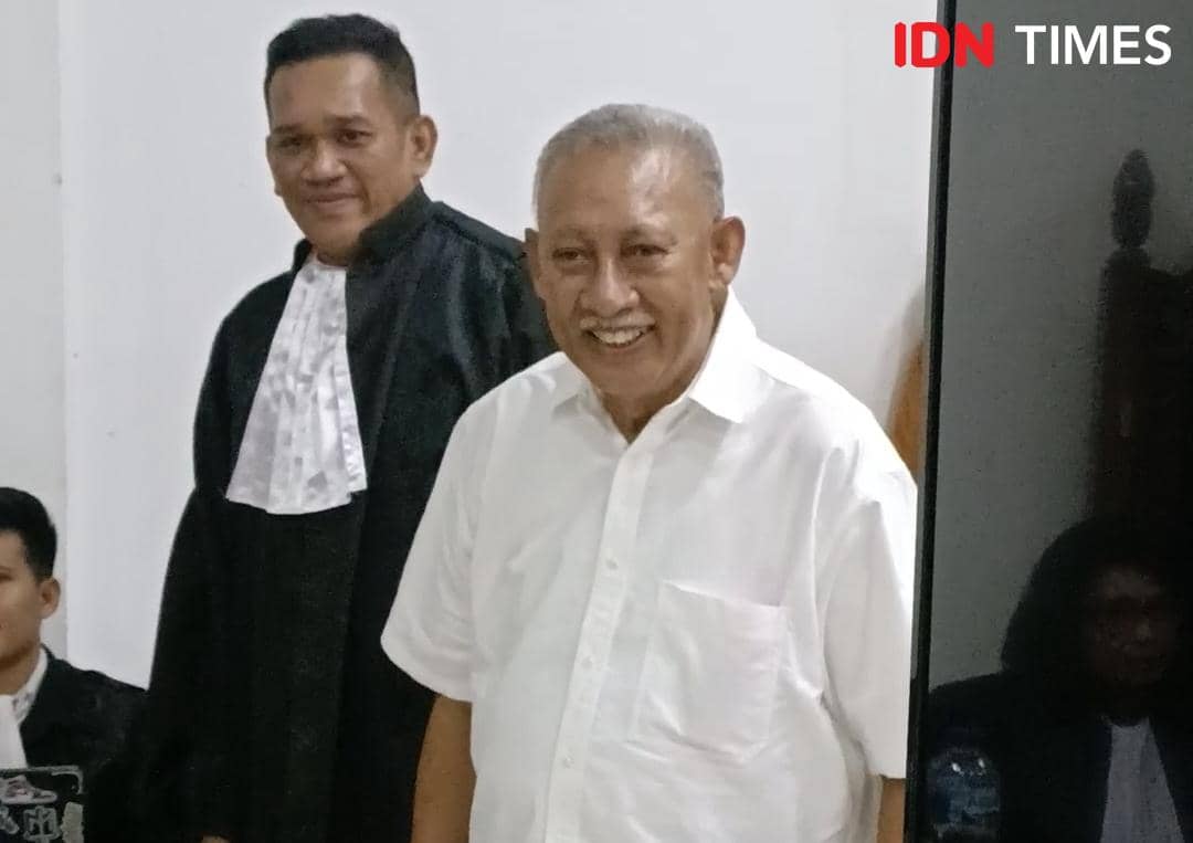 Seorang pria berusia lanjut mengenakan kemeja putih tersenyum di ruang sidang bersama seorang pria lain berpakaian toga hitam.