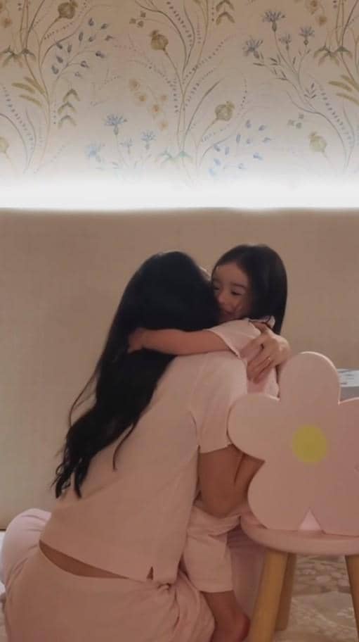 Seorang wanita berambut panjang mengenakan piyama berwarna lembut memeluk seorang anak kecil di ruangan dengan dekorasi bunga.