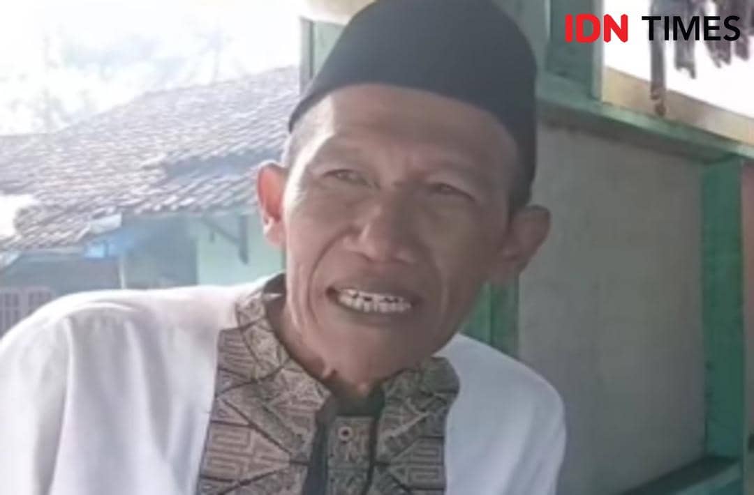 Seorang pria mengenakan peci hitam dan baju koko putih sedang berbicara di area rumah dengan latar bangunan beratap genteng.