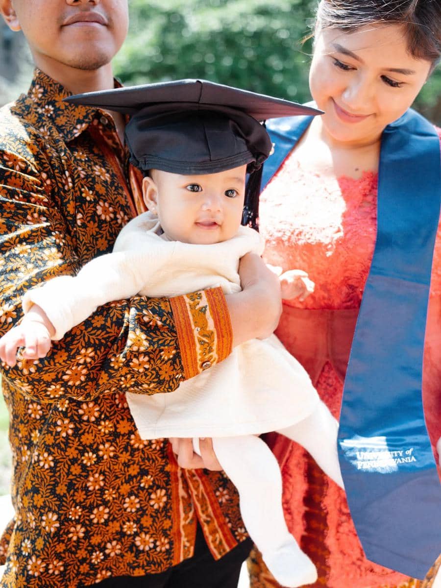 Seorang perempuan mengenakan selempang wisuda biru bersama pria berbaju batik, menggendong bayi memakai topi wisuda di luar ruangan.
