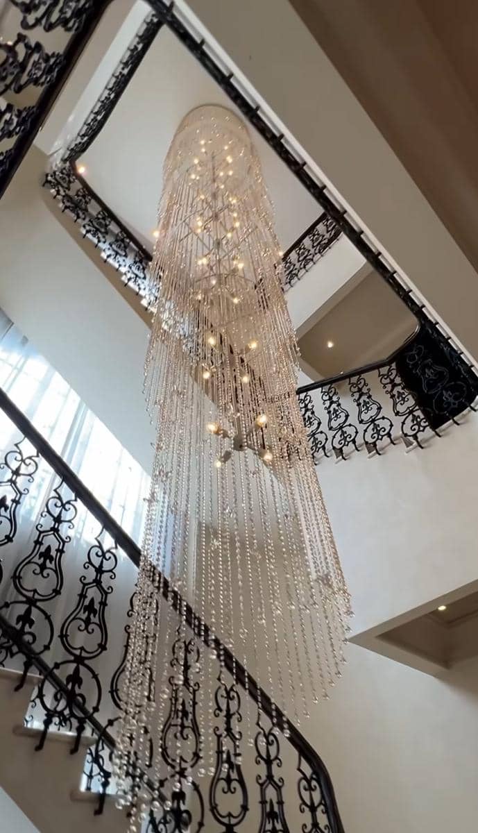 Lampu gantung kristal besar menggantung di tengah tangga melingkar dengan pagar besi berukir hitam di rumah mewah bertingkat.