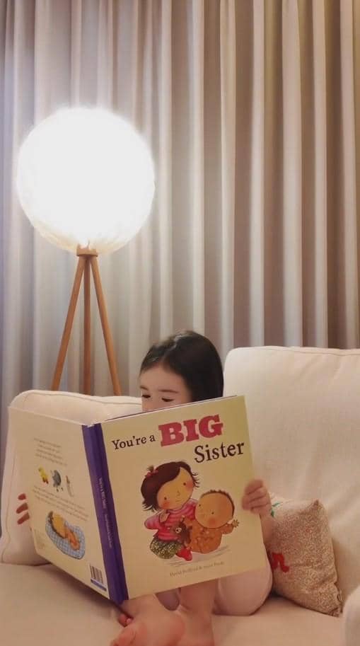 Seorang anak kecil duduk di sofa membaca buku bergambar berjudul 'You're a Big Sister' di ruang tamu dengan lampu berdiri menyala.