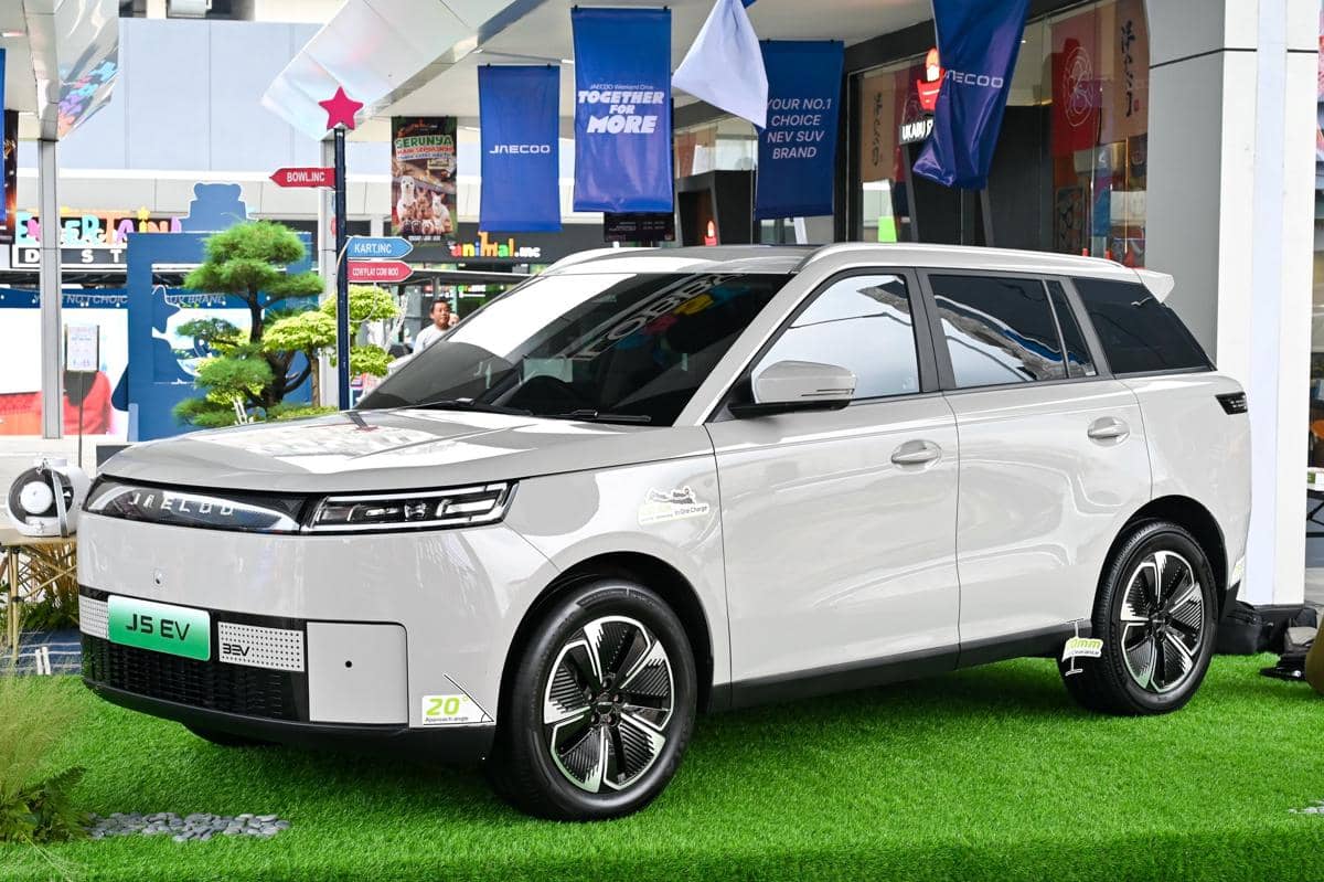 Mobil listrik Jaecoo J5 EV berwarna putih dipamerkan di area pameran dengan latar spanduk promosi dan dekorasi hijau.