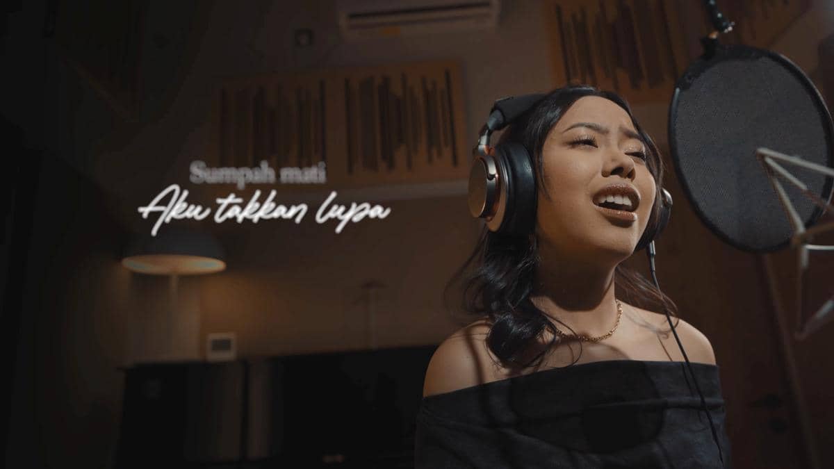 Seorang penyanyi perempuan mengenakan headphone sedang bernyanyi di depan mikrofon di studio rekaman dengan pencahayaan hangat.