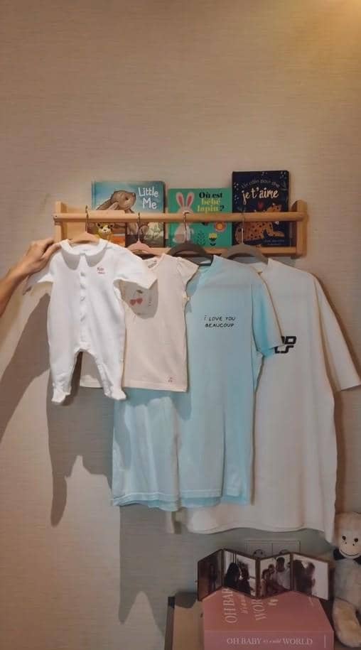 Deretan pakaian bayi dan dewasa berwarna putih dan biru muda tergantung di gantungan kayu dengan beberapa buku anak di atasnya.