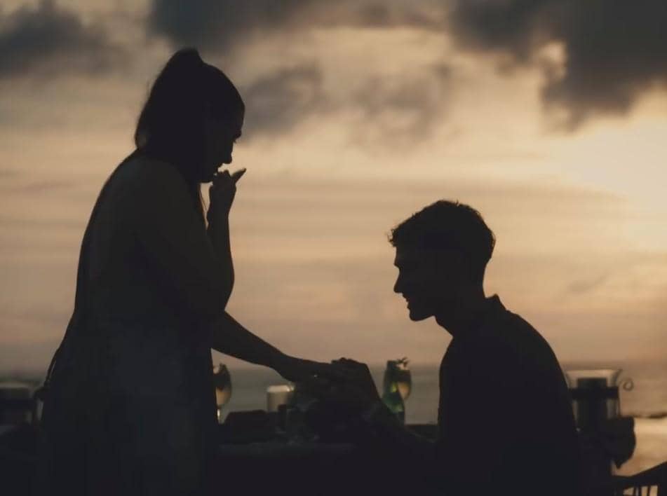 Seorang pria berlutut melamar kekasihnya di tepi pantai saat matahari terbenam dengan suasana romantis dan siluet indah.
