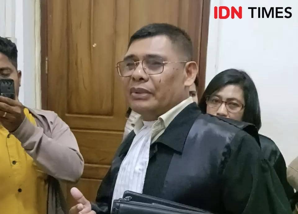 Seorang pria mengenakan jas hitam dan kemeja putih berbicara di depan pintu kayu, dikelilingi beberapa orang di ruang dalam gedung.