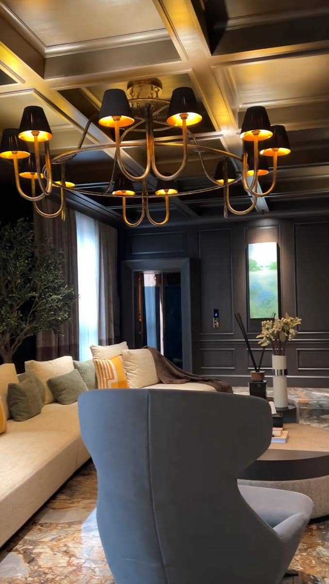 Ruang tamu bergaya modern dengan lampu gantung emas, sofa krem dengan bantal warna-warni, dan dinding panel gelap elegan.