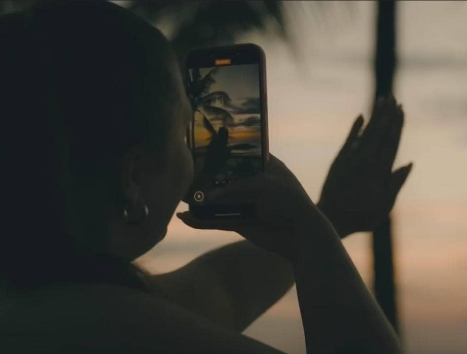 Seorang perempuan memotret pemandangan matahari terbenam di pantai dengan ponsel, menampilkan siluet pohon kelapa dan langit senja.