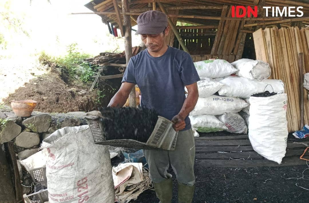 Seorang pria mengenakan topi dan kaus biru sedang menuangkan arang dari wadah di area produksi dengan tumpukan karung di sekitarnya.