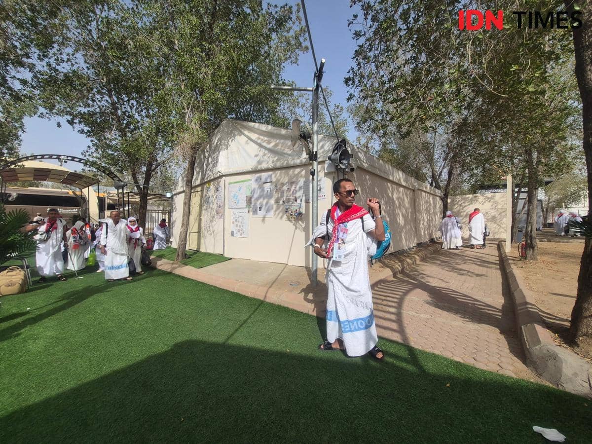 Jemaah haji asal Kalimantan Selatan mengenakan pakaian ihram dan berjalan menuju area tenda di Arafah di bawah pepohonan rindang.