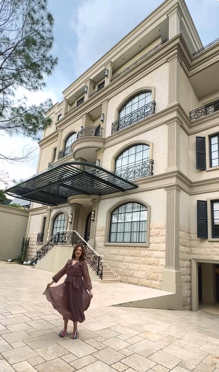 Seorang wanita berpose di halaman depan rumah besar bergaya klasik dengan dinding krem, balkon besi hitam, dan tangga elegan.