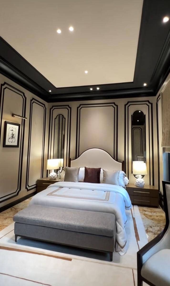 Kamar tidur mewah dengan desain klasik modern, tempat tidur besar berwarna netral, lampu meja di sisi kanan kiri, dan pencahayaan hangat.