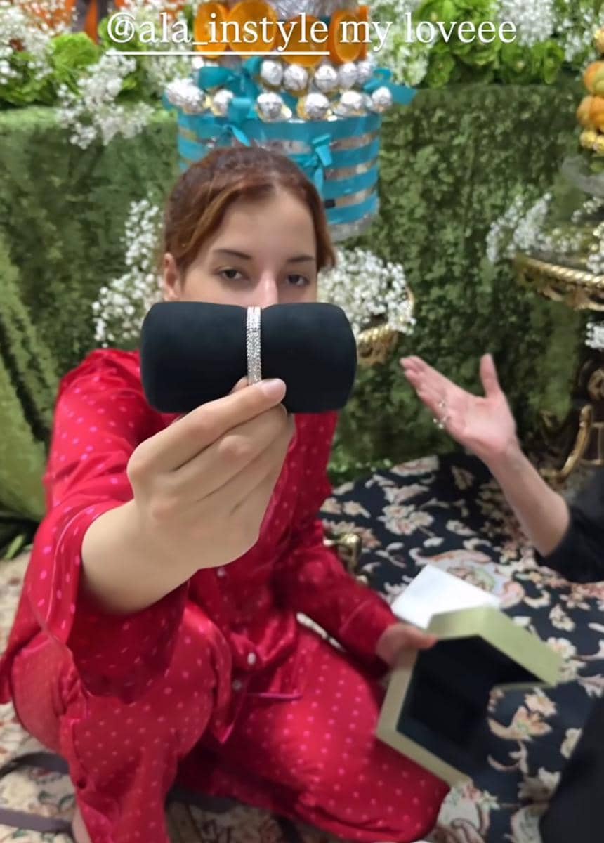 Seorang perempuan mengenakan piyama merah memegang kotak perhiasan hitam dengan latar dekorasi bunga dan hadiah ulang tahun.