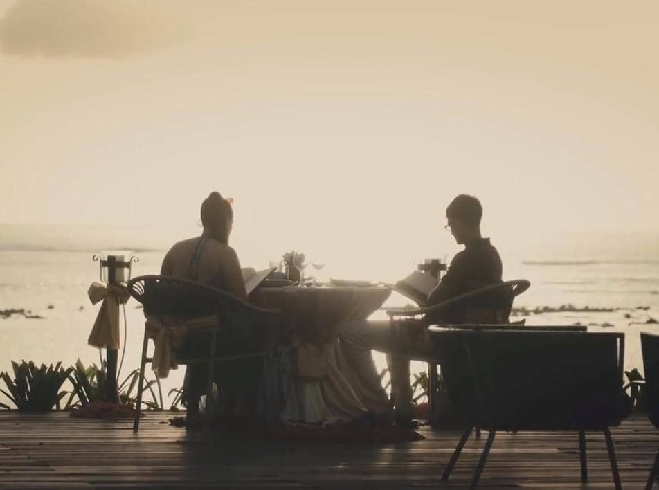Sepasang kekasih duduk di meja makan tepi pantai saat matahari terbenam, menikmati suasana romantis dengan pemandangan laut.