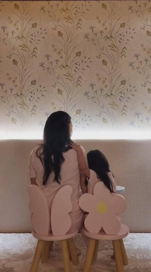 Seorang wanita berambut panjang duduk berdampingan dengan anak kecil di kursi berbentuk sayap dan bunga berwarna pink di ruangan bernuansa lembut.
