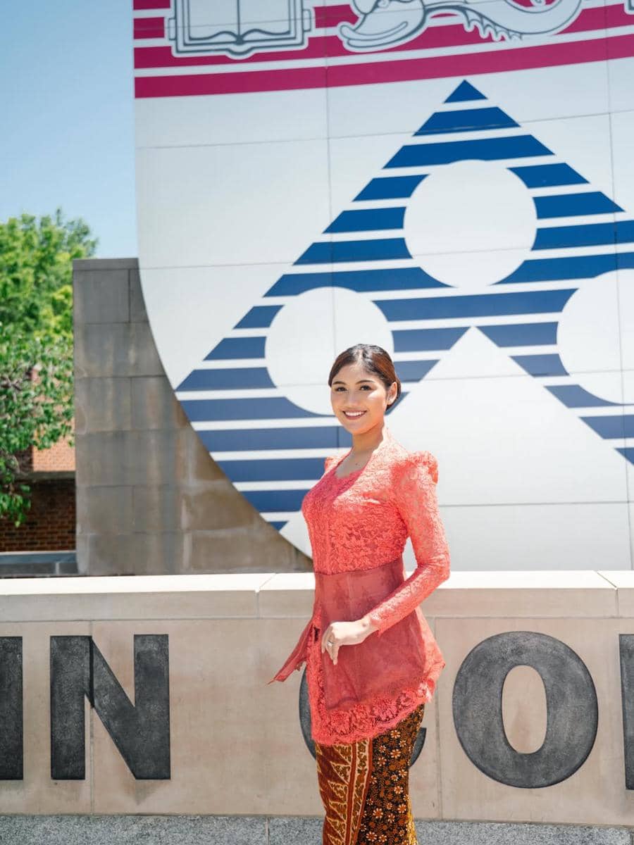 Seorang perempuan mengenakan kebaya merah dan kain batik berpose di depan dinding bertuliskan huruf besar di area kampus luar negeri.