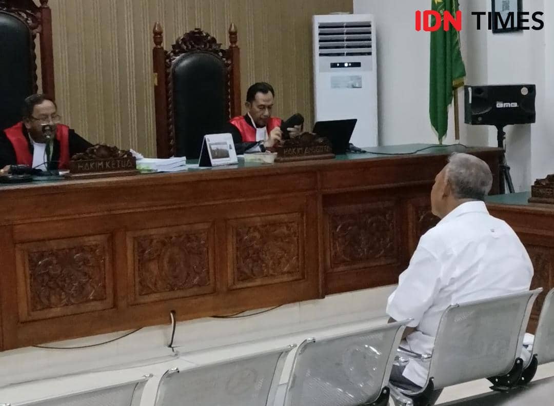 Suasana sidang di pengadilan dengan dua hakim mengenakan toga merah hitam dan seorang terdakwa duduk di kursi pengunjung mengenakan kemeja putih.