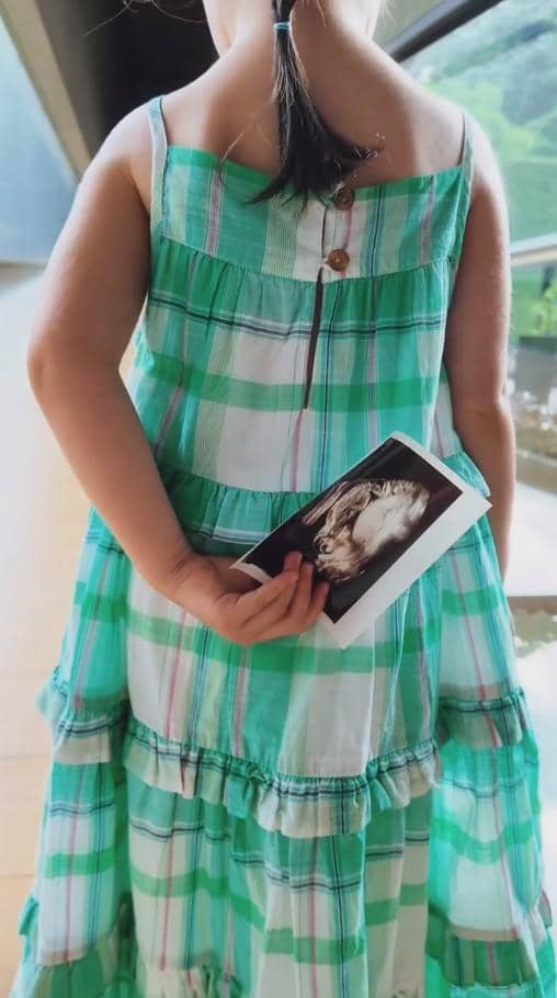 Seorang anak kecil mengenakan gaun kotak-kotak hijau putih memegang foto hasil USG di belakang punggungnya di dekat jendela besar.