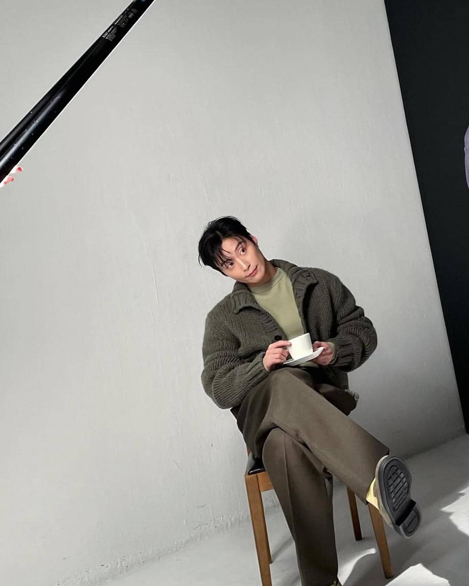 Seorang pria duduk santai di kursi sambil memegang cangkir, mengenakan sweater rajut abu-abu dan celana panjang cokelat dalam pencahayaan studio.
