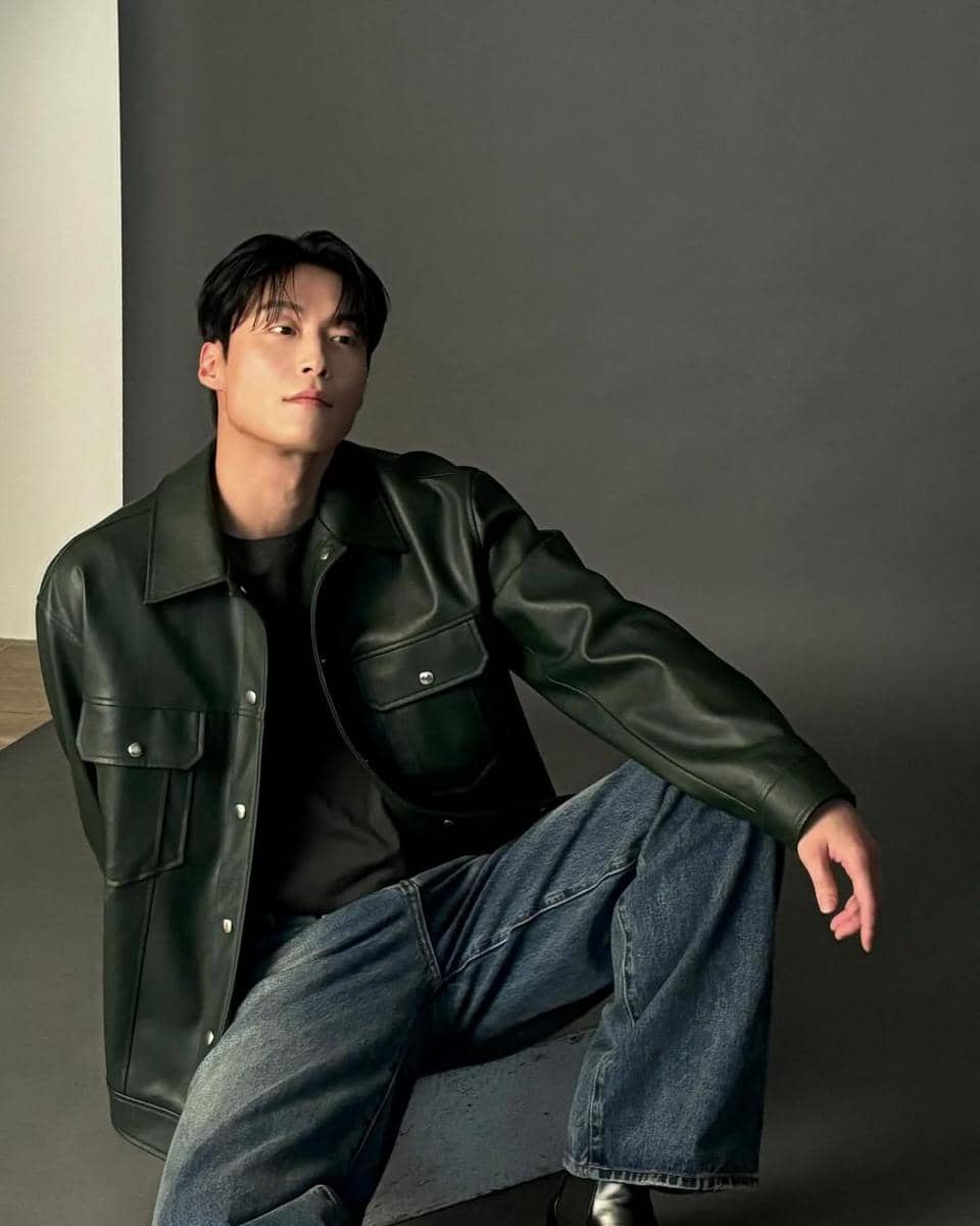 Seorang pria mengenakan jaket kulit hitam dan celana jeans biru duduk santai di studio dengan latar abu-abu gelap.