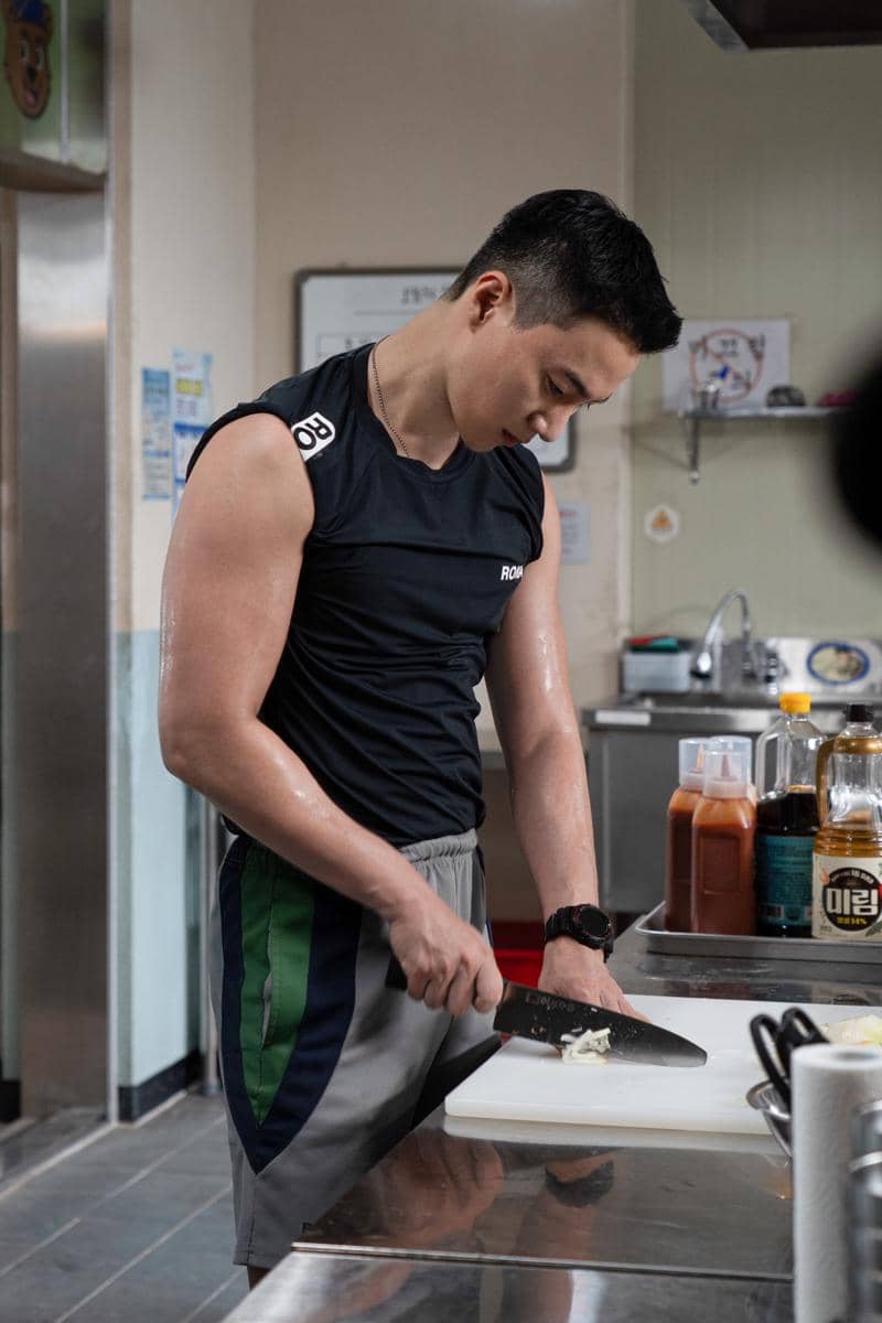 Seorang pria mengenakan kaus tanpa lengan hitam sedang memotong bahan makanan di dapur dengan pisau di atas talenan putih.
