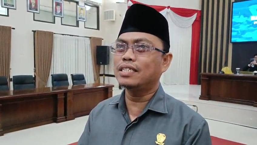 Seorang pria mengenakan peci hitam dan kemeja abu-abu berbicara di ruang rapat DPRD dengan meja kayu dan tirai putih di latar belakang.
