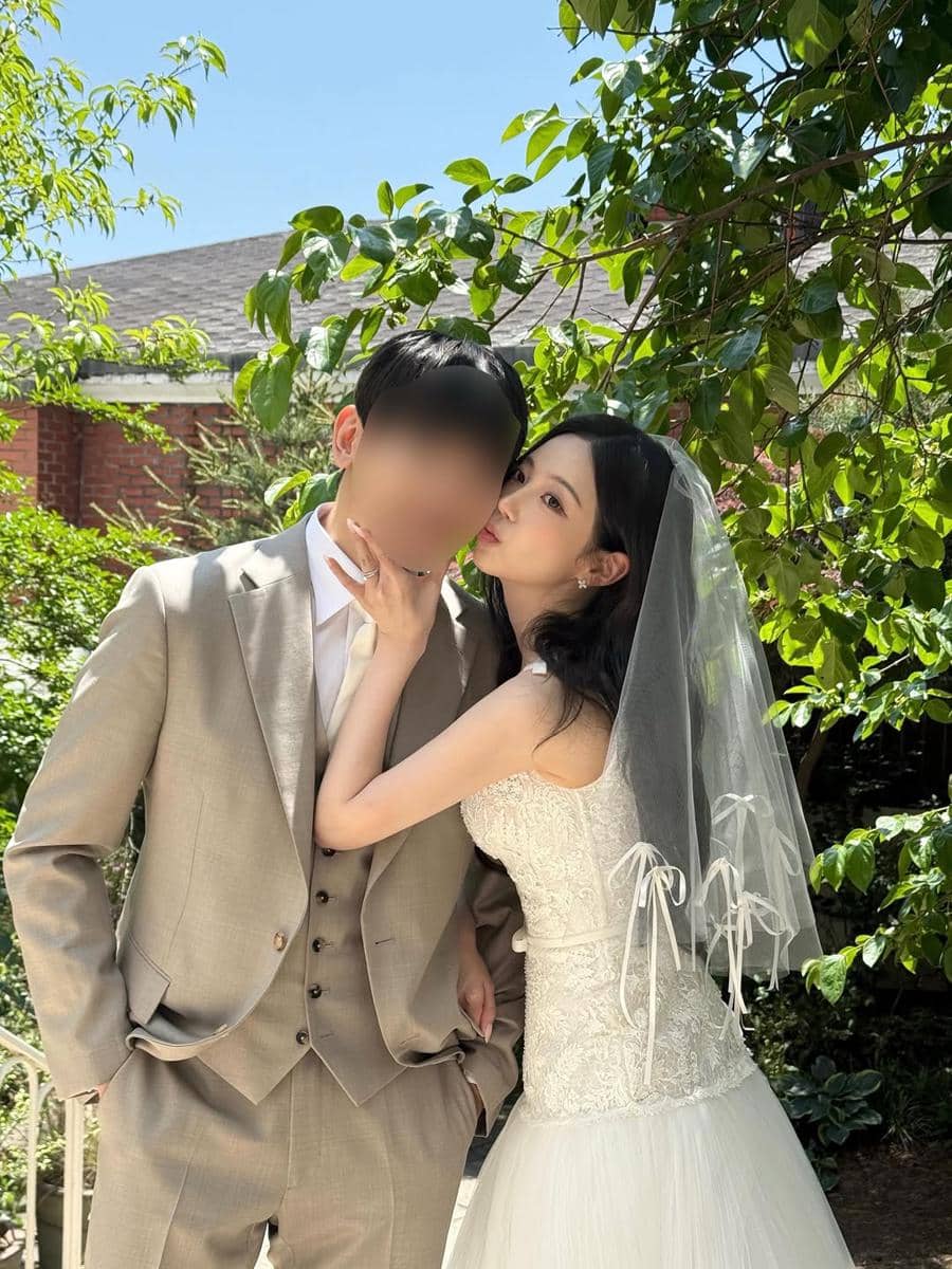 Seorang wanita mengenakan gaun pengantin putih dan veil berpose mesra dengan pria bersetelan jas krem di taman yang rindang.