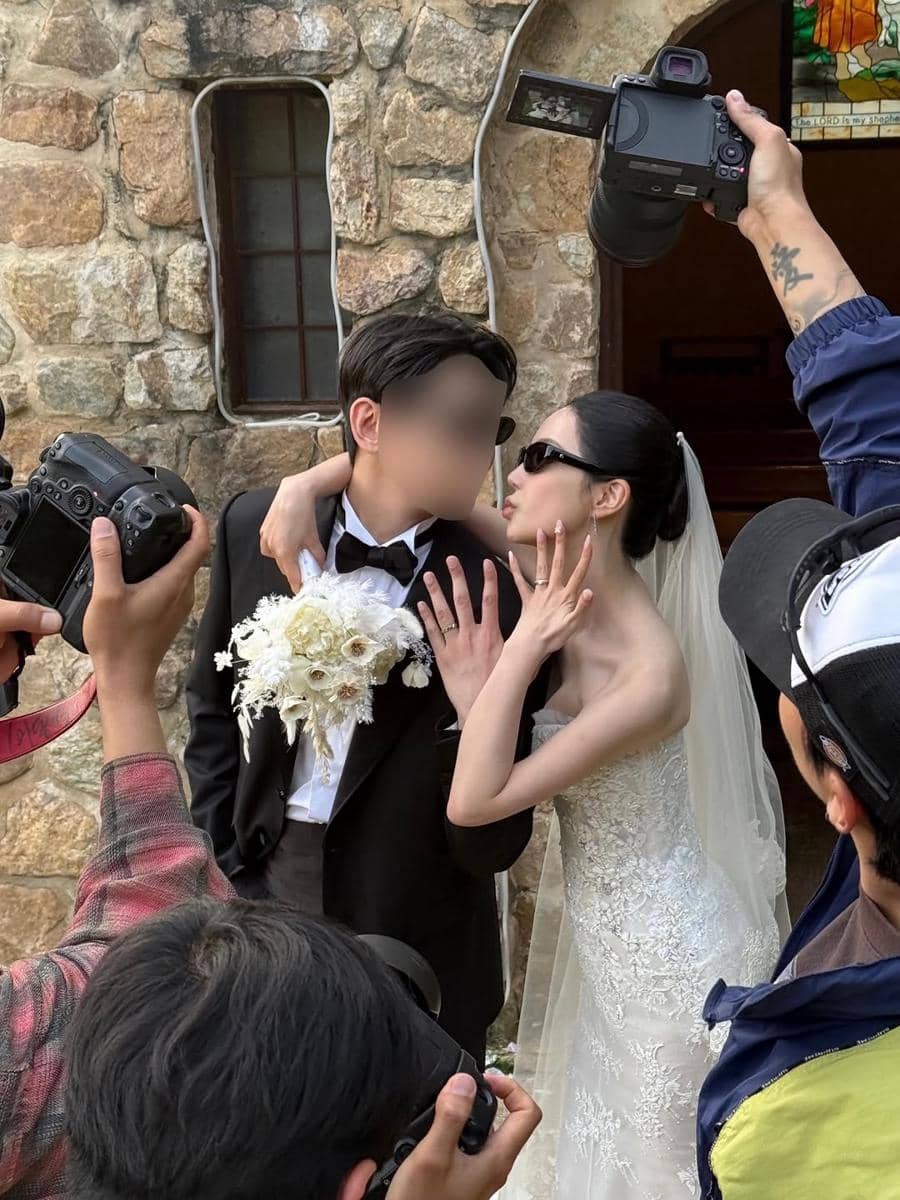 Pasangan mengenakan busana pengantin berpose mesra di depan bangunan batu sambil dikelilingi fotografer yang mengambil gambar.