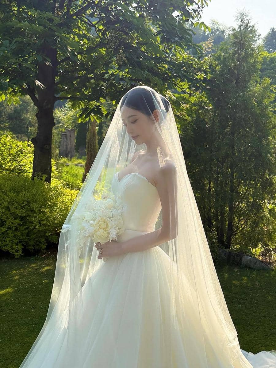 Seorang wanita mengenakan gaun pengantin putih dan veil panjang sambil memegang buket bunga di taman yang hijau dan teduh.