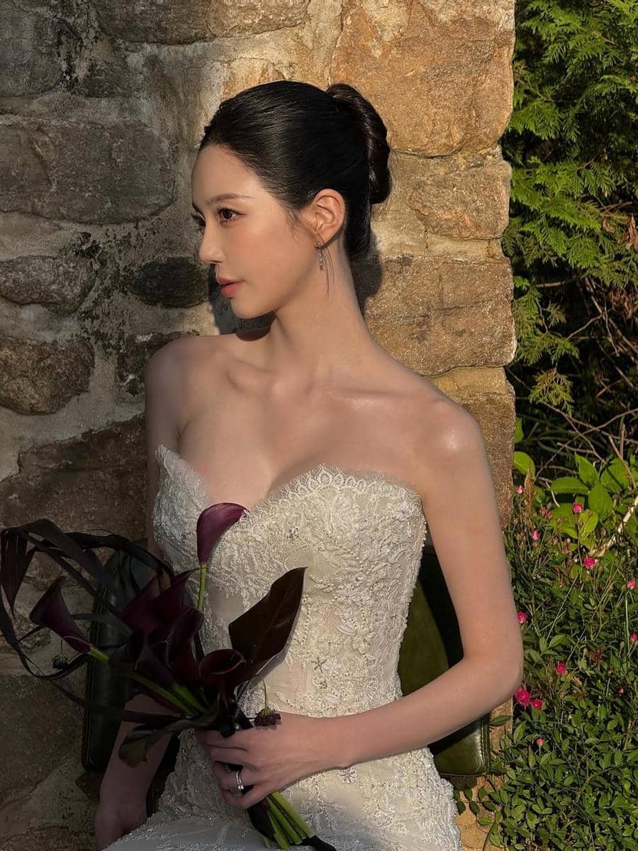 Seorang wanita mengenakan gaun pengantin putih berenda sambil memegang buket bunga ungu gelap di dekat dinding batu dan tanaman hijau.
