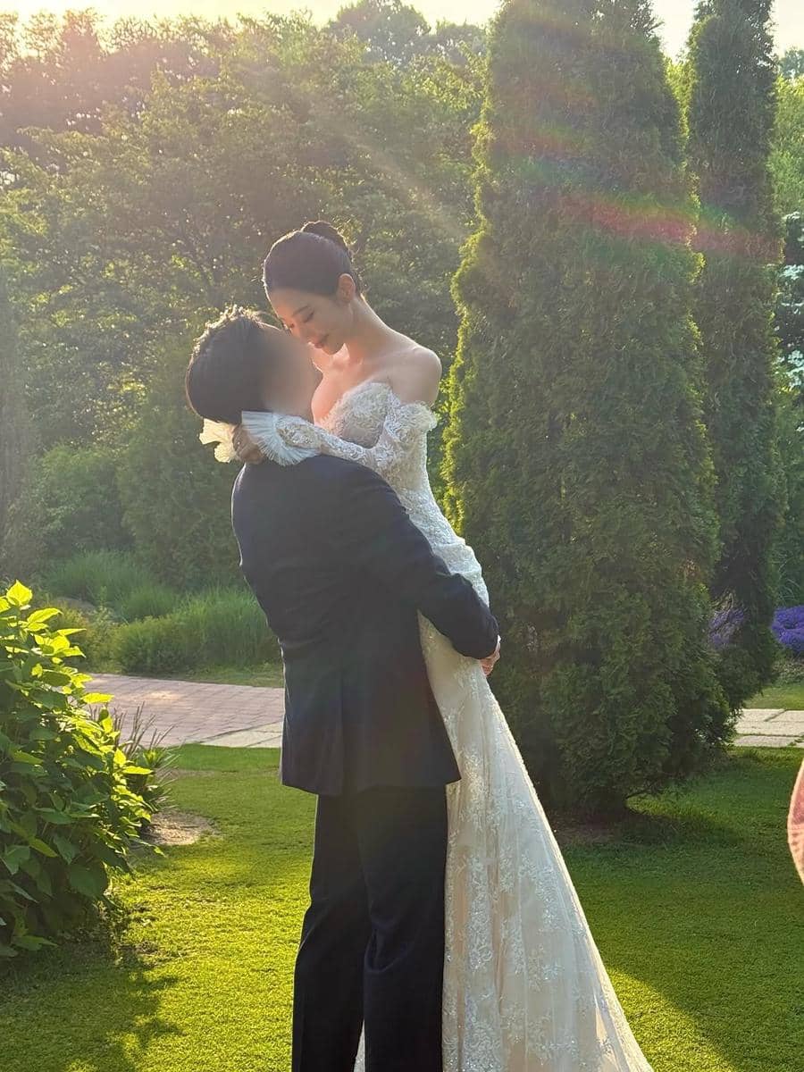 Pasangan mengenakan gaun pengantin dan setelan jas berpose romantis di taman hijau dengan cahaya matahari sore yang hangat.