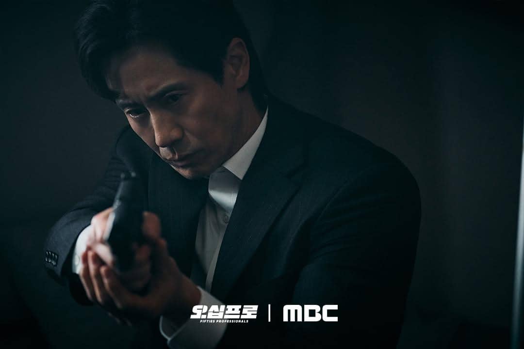Seorang pria berjas hitam memegang pistol dengan ekspresi serius dalam adegan drama Fifties Professionals produksi MBC.