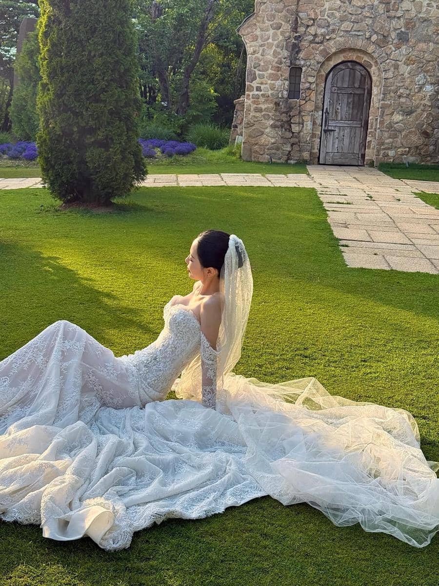 Seorang wanita mengenakan gaun pengantin putih dengan detail renda dan kerudung panjang duduk di taman hijau dekat bangunan batu.
