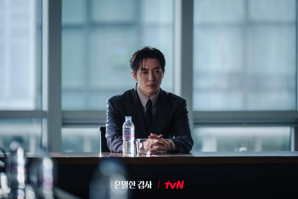 Seorang pria mengenakan setelan jas duduk di meja dengan botol air dan gelas di depannya dalam adegan drama Korea Filing for Love.