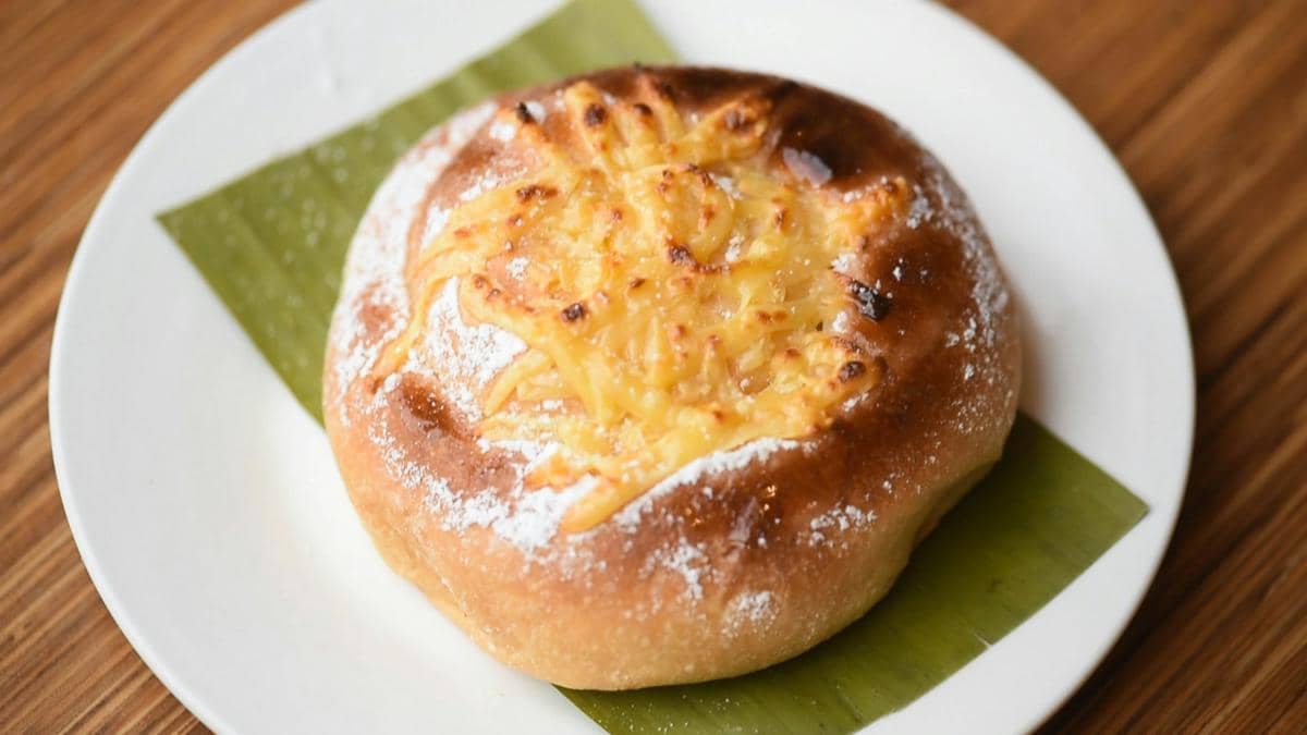 Donat berwarna keemasan dengan taburan keju parut dan gula halus di atas piring putih beralaskan daun pisang.