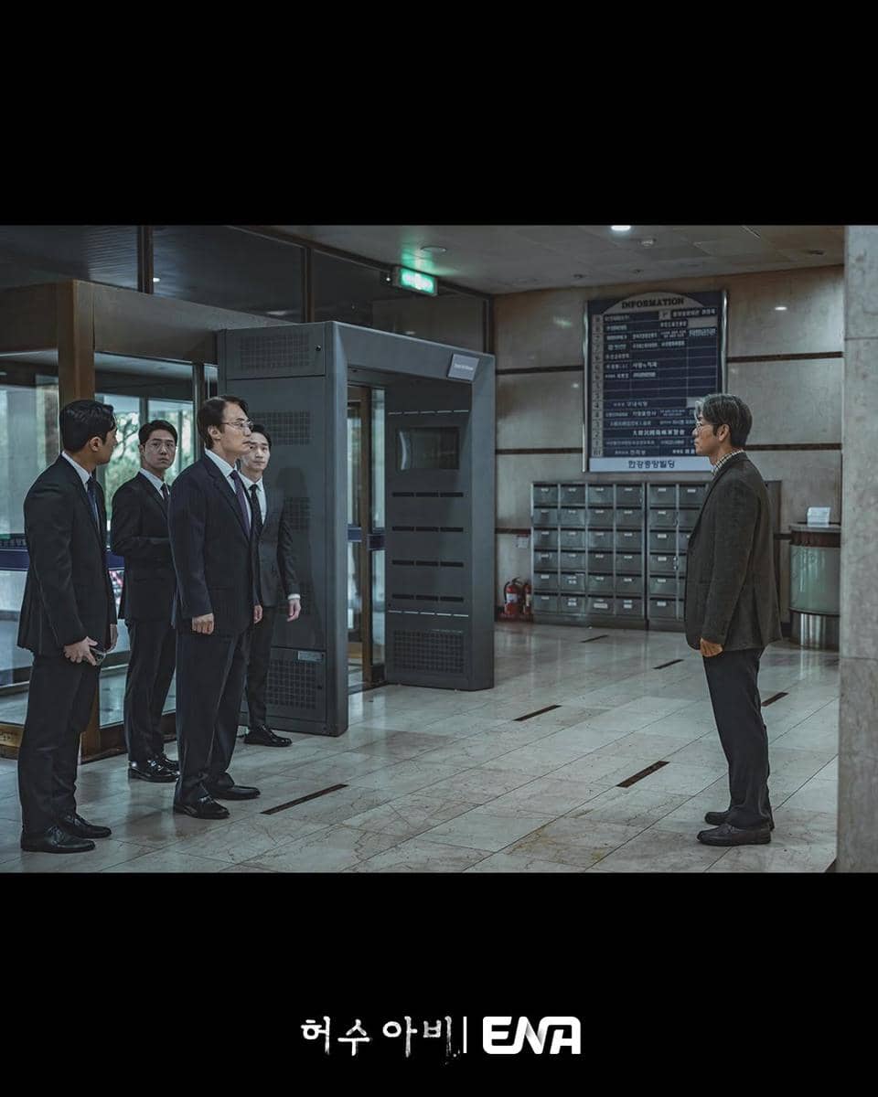 Beberapa pria berjas berdiri di dalam gedung kantor modern, berhadapan dengan seorang pria berjaket di depan mesin pemindai keamanan.
