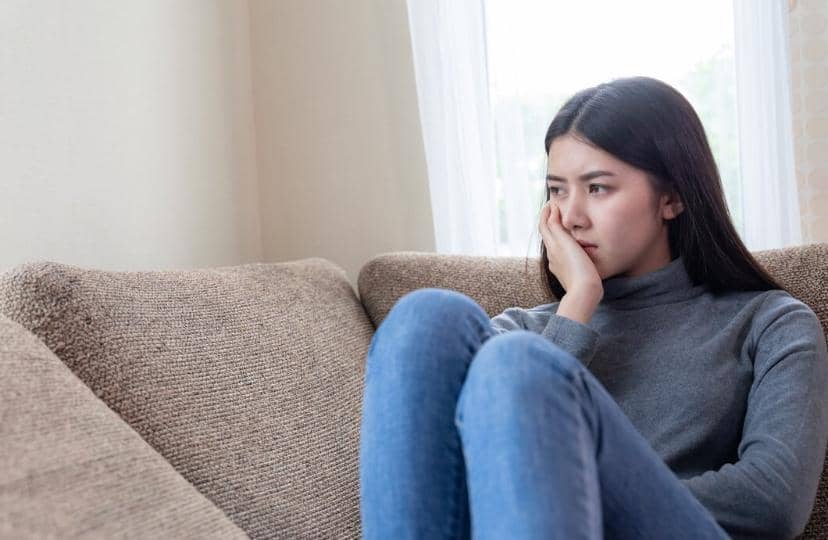 Seorang wanita duduk di sofa dengan ekspresi murung, mengenakan sweter abu-abu dan celana jeans, tampak termenung di dekat jendela.