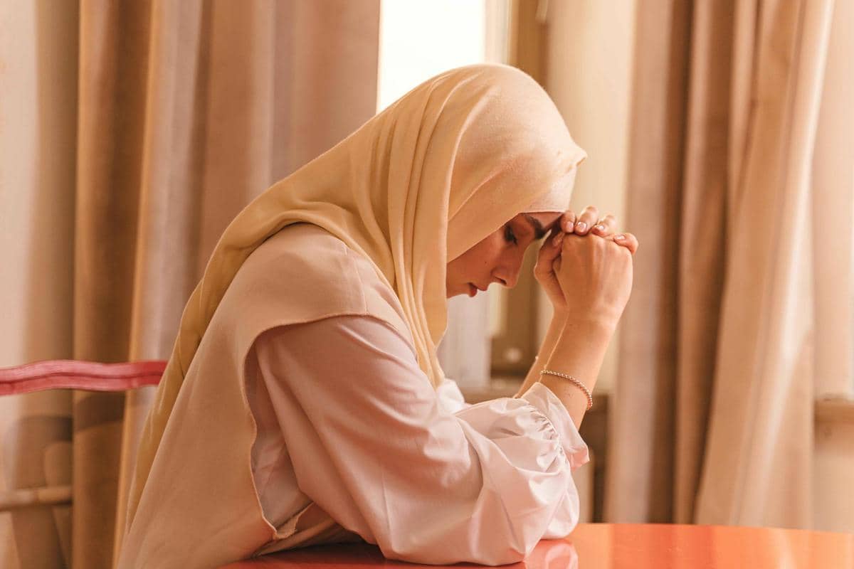 Seorang perempuan berhijab duduk dengan tangan bertumpu di dahi, tampak menenangkan diri di dalam ruangan bernuansa hangat.